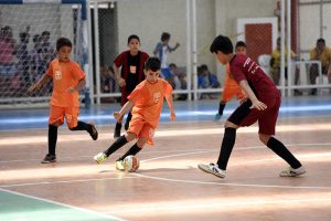 Futsal Sesc: a foto mostra cinco meninos jogando futsal em uma quadra, três com uniforme laranja e dois de uniforme vinho.