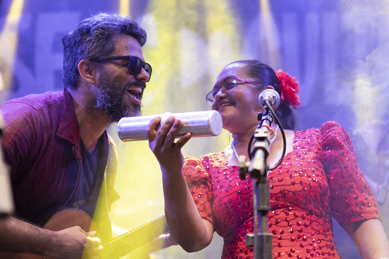 Mostra Cariri: imagem mostra um homem e uma mulher cantando juntos em um palco bem iluminado, em plano fechado.