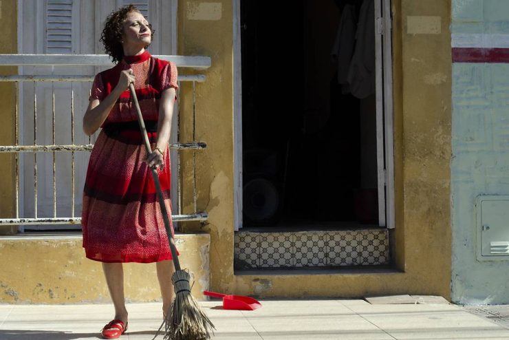 Dia da Mulher: foto mostra Pacarrete, personagem principal do filme homônimo, em frente a uma casa com vestido vermelho e vassoura na mão. Ela olha para cima em pose altiva. 