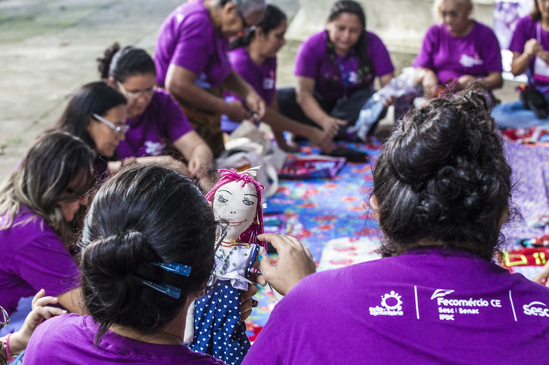 Projetos comunitários: na foto, há uma roda de mulheres com camisas roxas sentadas no chão em cima de um tecido colorido. Algumas delas tem bonecas de pano na mão.