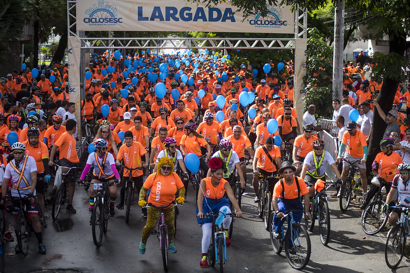 Passeio ciclístico CicloSesc: imagem mostra vários ciclistas saindo da faixa de largada com camisas laranjas e balões azuis.