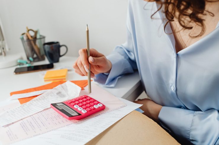 Para reduzir custos: a imagem mostra parte do corpo de uma mulher com uma camisa social e segurando um lápis enquanto faz contas em uma calculadora. Ao lado da calculadora, em uma mesa, há várias notas fiscais e papéis. 
