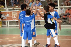 Futsal Sesc: na imagem, cinco meninos dão um "high five" em uma quadra de futsal. Todos usam uniforme azul do Sesc, meiões brancos e chuteira.