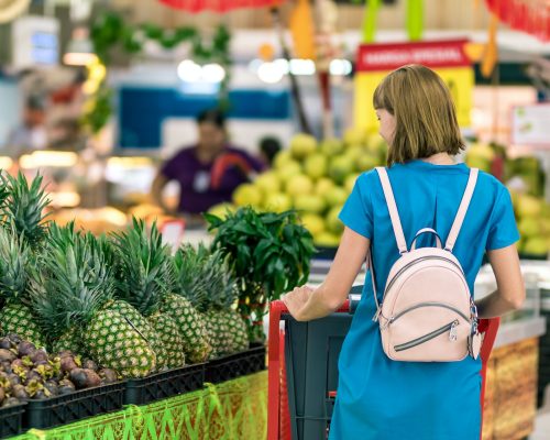 Pesquisa da Fecomércio: Uma mulher faz compras em um supermercado. Ela é branca, usa cabelo loiro e curto e um vestido azul e mochila. Está de costas para a câmera observando uma prateleira com abacaxis.