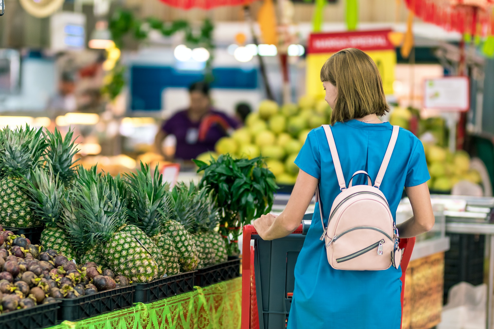 Pesquisa da Fecomércio: Uma mulher faz compras em um supermercado. Ela é branca, usa cabelo loiro e curto e um vestido azul e mochila. Está de costas para a câmera observando uma prateleira com abacaxis.