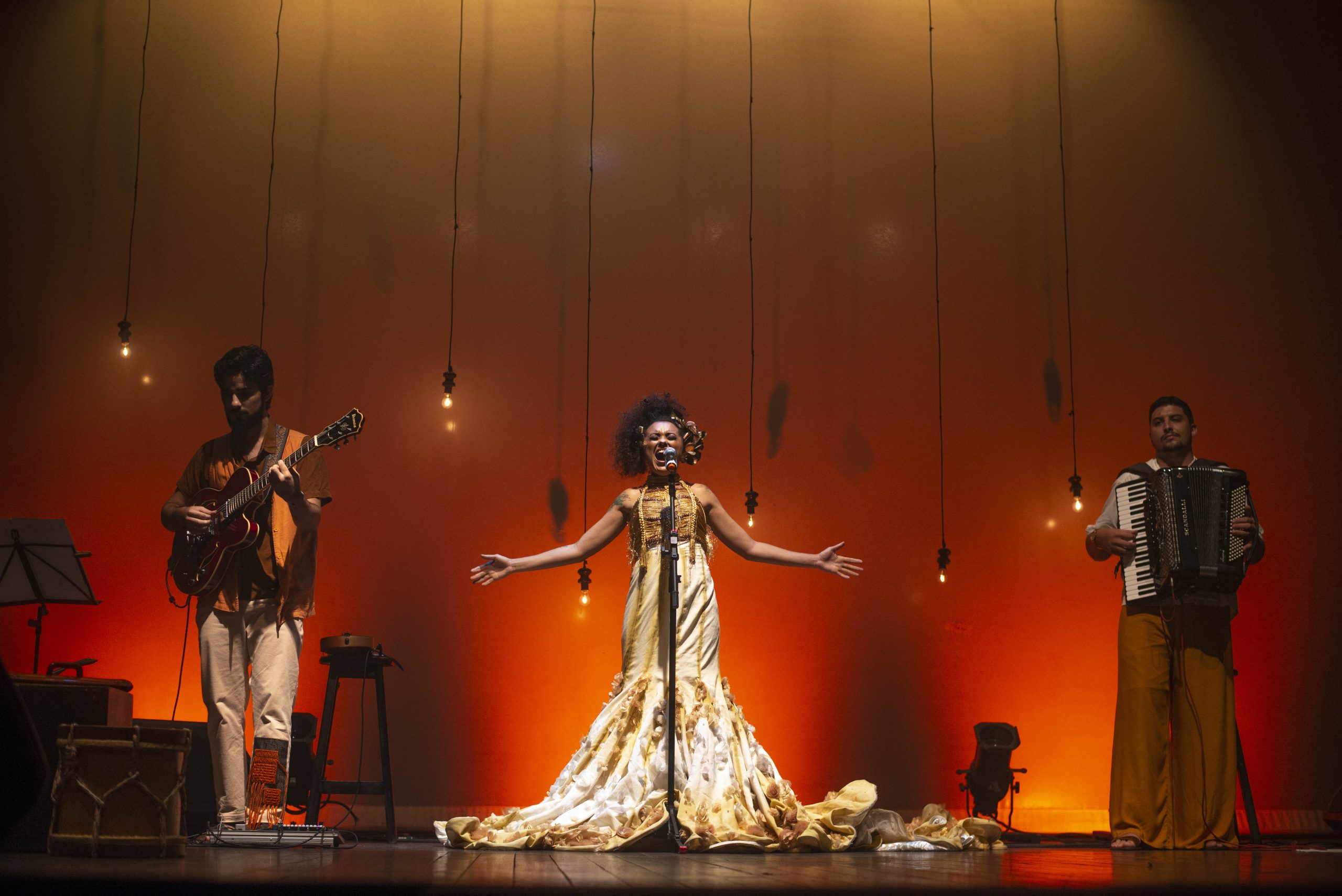 Programação Sesc: foto da cantora Luana Florentino em um palco bem iluminado, nas cores vermelho e laranja. Ela é uma mulher negra, de cabelo cacheado e preto, e usa um vestido longo e claro enquanto levanta as mãos para o alto. Também há um músico de cada lado do palco.