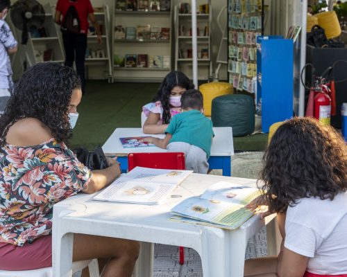 Programação Sesc: a imagem mostra algumas mesinhas de plástico com livros infantis. Ao redor das mesas, duas mulheres adultas com máscaras de proteção leem junto a um menino e uma menina.
