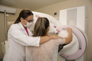 Maracanaú: na imagem, uma mulher auxilia outra a fazer a mamografia. A paciente está de costas e se apoia na máquina para realizar o procedimento. Ambas são brancas e jovens, têm cabelo claro e liso e estão paramentadas.