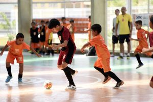 Futsal no Sesc: foto mostra quatro meninos jogando futsal em uma quadra. Três deles usam camisa de mesma cor, laranja, e o quarto um uniforme vinho. No fundo da imagem, pode-se ver um treinador e mais alunos.