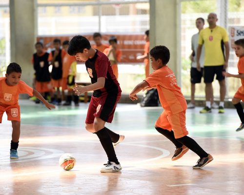 Futsal no Sesc: foto mostra quatro meninos jogando futsal em uma quadra. Três deles usam camisa de mesma cor, laranja, e o quarto um uniforme vinho. No fundo da imagem, pode-se ver um treinador e mais alunos.
