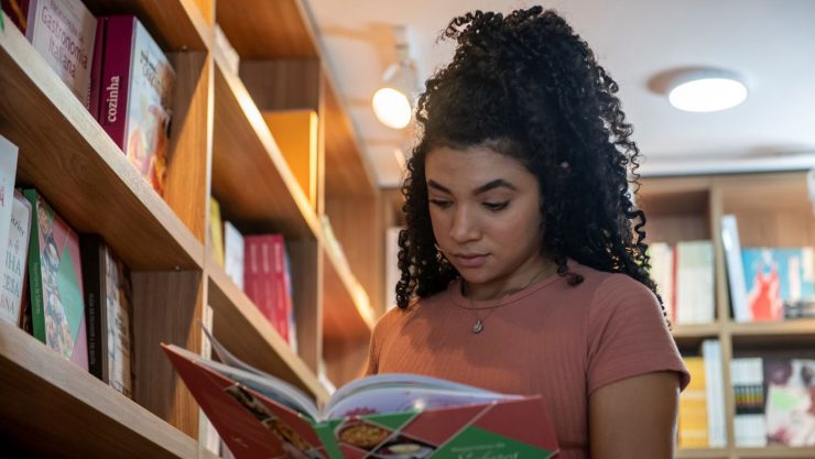 Bazar de São João: uma mulher jovem folheia um livro ao lado de uma estante da Livraria Senac. A moça usa uma camiseta marrom, tem pele negra, cabelo preto e cacheado preso em um coque e usa um colar.