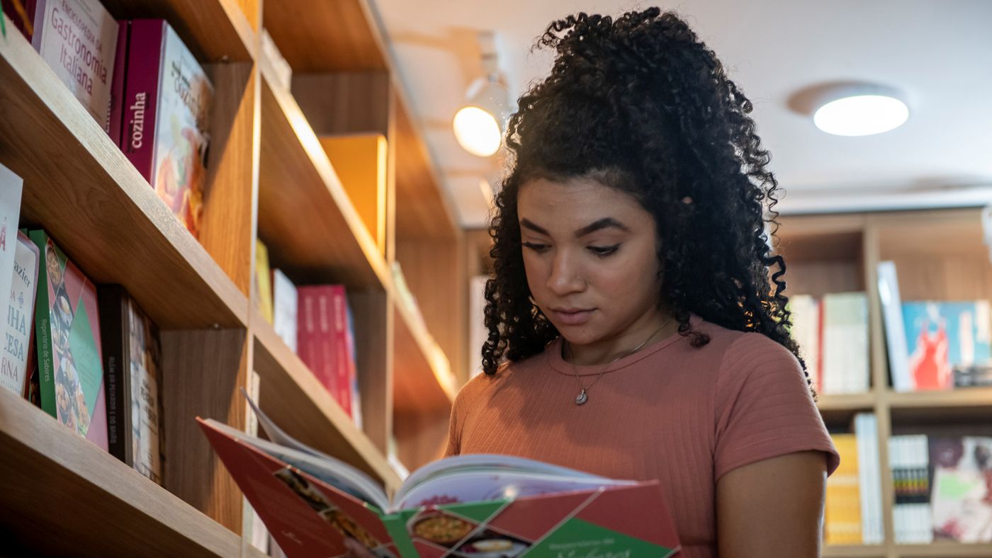 Bazar de São João: uma mulher jovem folheia um livro ao lado de uma estante da Livraria Senac. A moça usa uma camiseta marrom, tem pele negra, cabelo preto e cacheado preso em um coque e usa um colar.