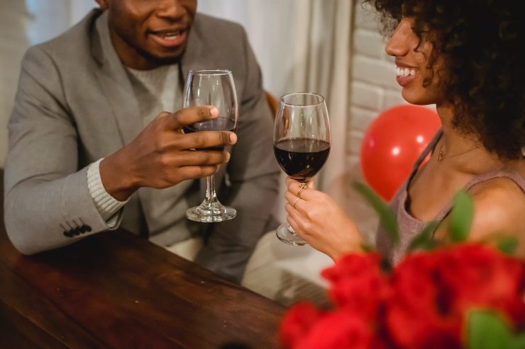 Dia dos Namorados: a foto mostra parcialmente um homem e uma mulher com trajes sociais, brindando taças de vinho e sorrindo um para o outro. Ambos são negros e a mulher tem cabelos cacheados