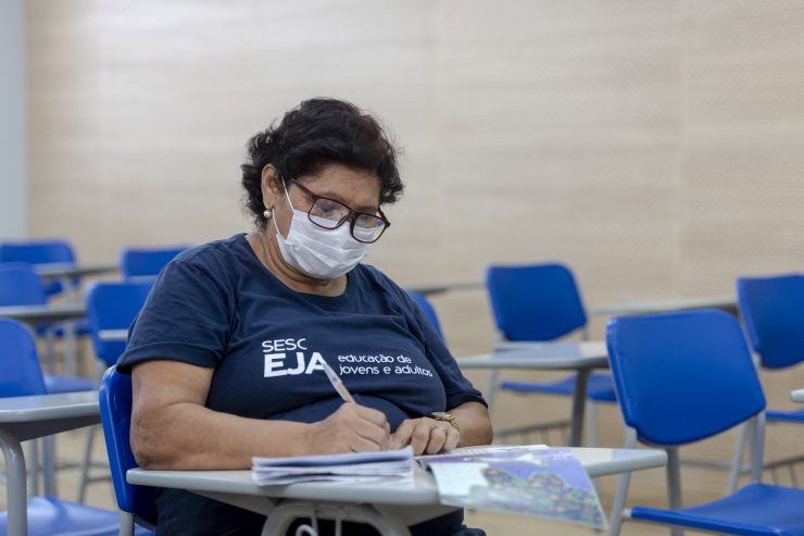 Educação de Jovens e Adultos: a imagem mostra uma mulher em uma sala de aula. Ela está sentada em uma carteira azul, usa óculos, máscara de proteção e camisa SESC EJA, enquanto escreve em um caderno