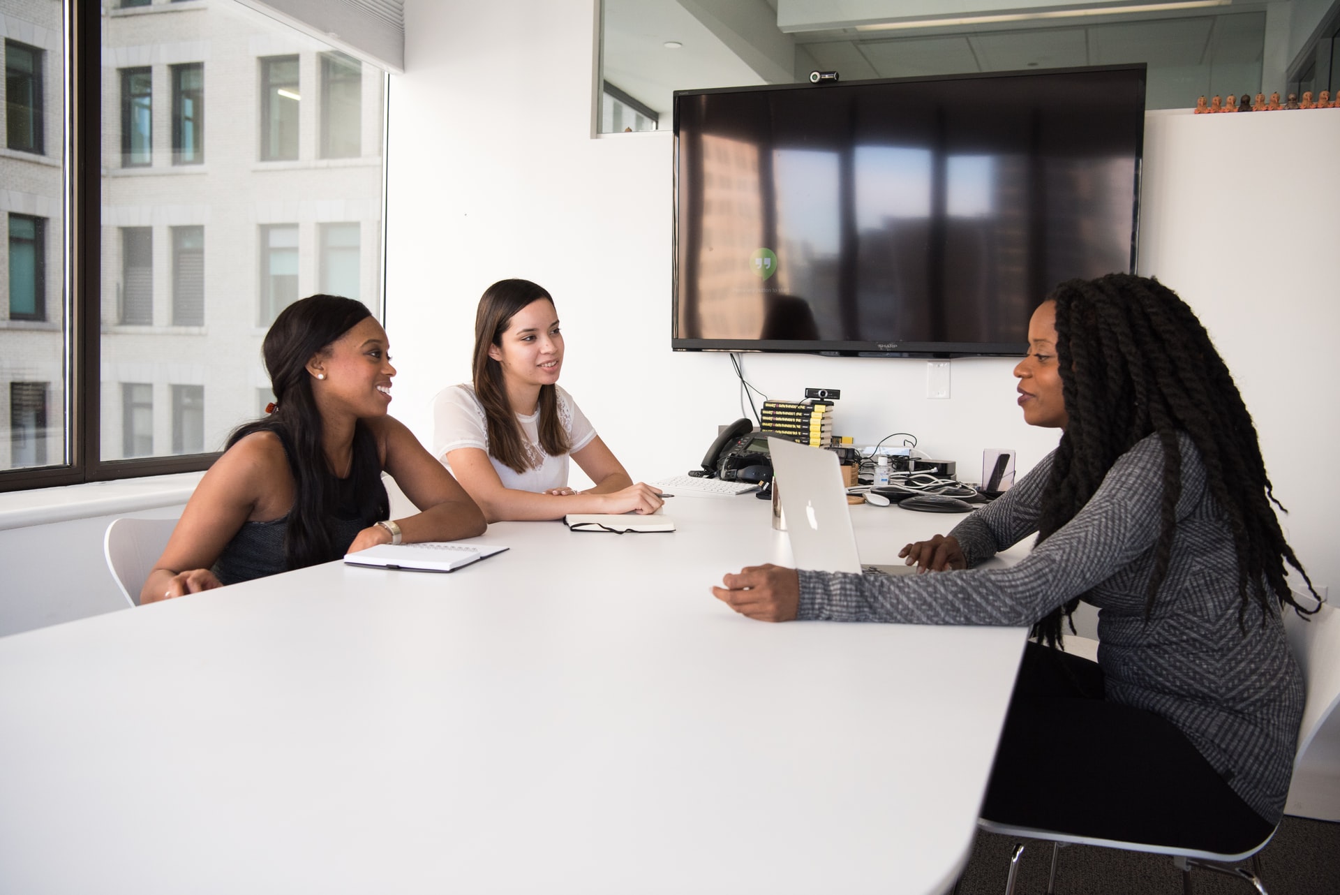 Empreendedorismo feminino: a imagem mostra uma mesa de reunião branca em frente a uma TV. Sentadas em frente a ela estão três jovens mulheres, duas negras e uma branca, conversando enquanto digitam em notebooks e escrevem em cadernos.
